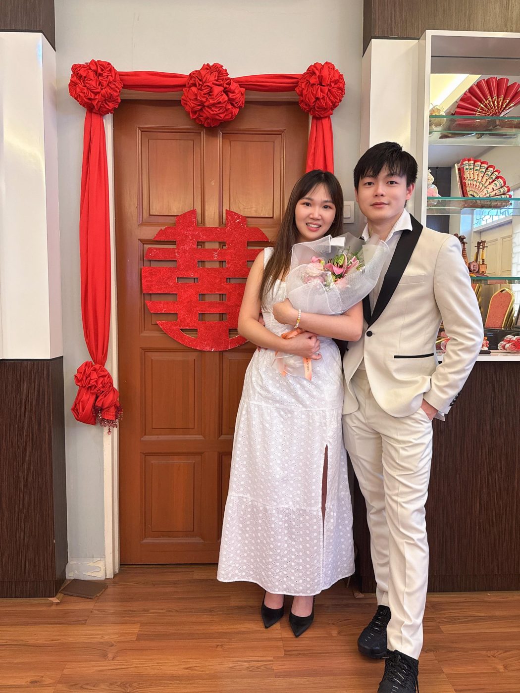 Yi Lin and Yu En in white, holding a bouquet in front of a red double-happiness door